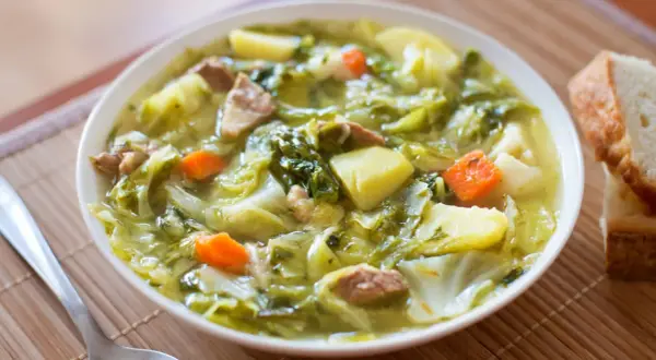 Bol de soupe au chou vert, pommes de terre, carottes et viande, avec du pain à côté.